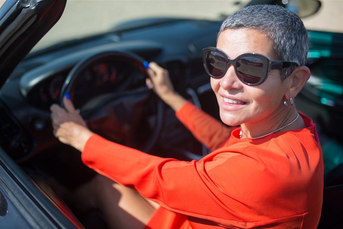 A smiling driver with short grey hair and sunglasses, both hands on the wheel of her car.