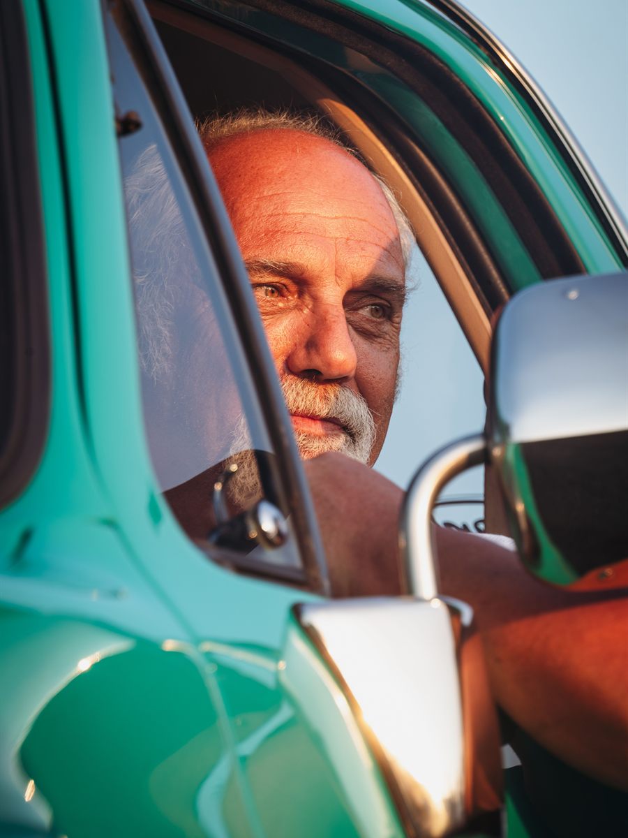 A grey-haired driver at the wheel of his car in late afternoon light, looking ahead down the road.